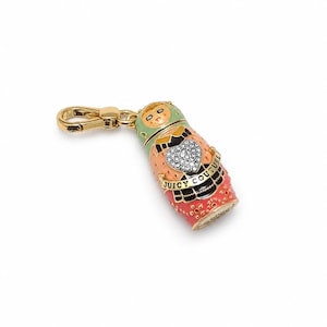 May include: A gold-tone metal charm shaped like a Russian nesting doll with enamel detailing in shades of green, peach, and red. The charm features a central heart encrusted with clear crystals and a banner with the text Juicy Couture.