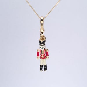 May include: Gold-tone necklace with a charm shaped like a toy soldier. The soldier is painted in red, white, black, and gold, with a black hat and a stern expression. The necklace chain is delicate.