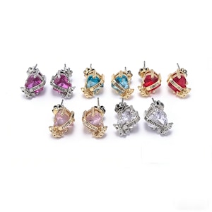 May include: A collection of Juicy Couture heart-shaped stud earrings in various colors, including pink, red, blue, and clear. Each earring features a crown design and the brand name. The earrings are set against a white background.
