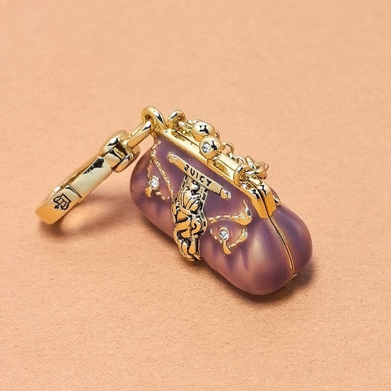 May include: A gold-coloured charm in the shape of a small, purple handbag with the word "JUICY" on it. The charm has a gold clasp and is adorned with small, clear crystals. The charm is on a light brown background.