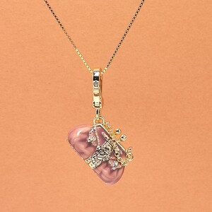 May include: A gold necklace with a pendant. The pendant has a pink, heart-shaped stone and a gold-coloured frame with a hand and crown design. The necklace hangs against a peach background.