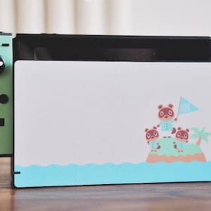 May include: A light blue and green Nintendo Switch console with a custom skin featuring characters from the Animal Crossing video game series. The skin shows Tom Nook and two villagers standing on an island with a palm tree and a blue ocean.