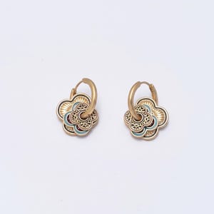 May include: A pair of gold-colored hoop earrings. Each earring features a four-petal flower design with intricate patterns and accents of teal and burgundy. The earrings have a vintage or bohemian aesthetic.