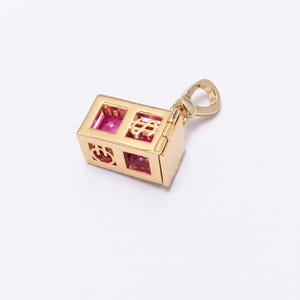 Pode incluir: Um berloque dourado em forma de casa pequena, com desenhos recortados e pedras preciosas rosa. O berloque tem uma porta articulada e uma argola para prender a um colar ou pulseira. O design da casa inclui um símbolo de dólar.