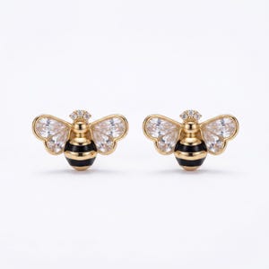 May include: A pair of gold-toned bee-shaped earrings. The earrings feature clear, faceted wings, a black and gold striped body, and a small cluster of clear stones on the head. The earrings are set against a white background.