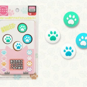 May include: A set of four paw-shaped controller caps for video game consoles. The caps are in various shades of blue and green with white paw prints. The caps are packaged in a clear plastic bag with a pink and white label that says "Super Mario Bros. 2" in Japanese.