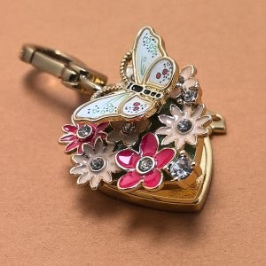 May include: A gold-coloured charm with a butterfly and floral design. The butterfly has white wings with blue and red accents. The flowers are pink and white, with clear crystal accents. The charm has a heart-shaped base and a clasp.