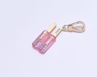 May include: A gold-coloured charm featuring two pink popsicle-shaped pendants with glitter accents. The charm has a gold-coloured clasp and the word "JUICY" is printed on one of the pendants.