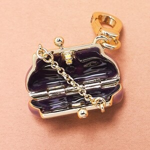 May include: A gold-coloured charm in the shape of a small, open purse. The interior is a deep purple, and a delicate gold chain hangs from the purse. The charm has a clasp for attaching to a bracelet or necklace.