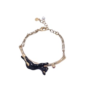 Pode incluir: Pulseira dourada com um design de gato preto. O gato está deitado numa barra dourada curva, com uma pequena gema rosa e um detalhe de pérola. A pulseira tem uma corrente delicada e um fecho de lagosta.