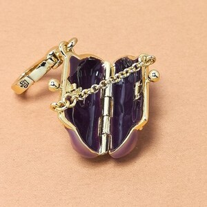 May include: A gold-coloured, hinged locket with a deep purple interior and a delicate chain. The locket is adorned with gold accents and a small, decorative clasp. The design resembles a miniature book or purse.