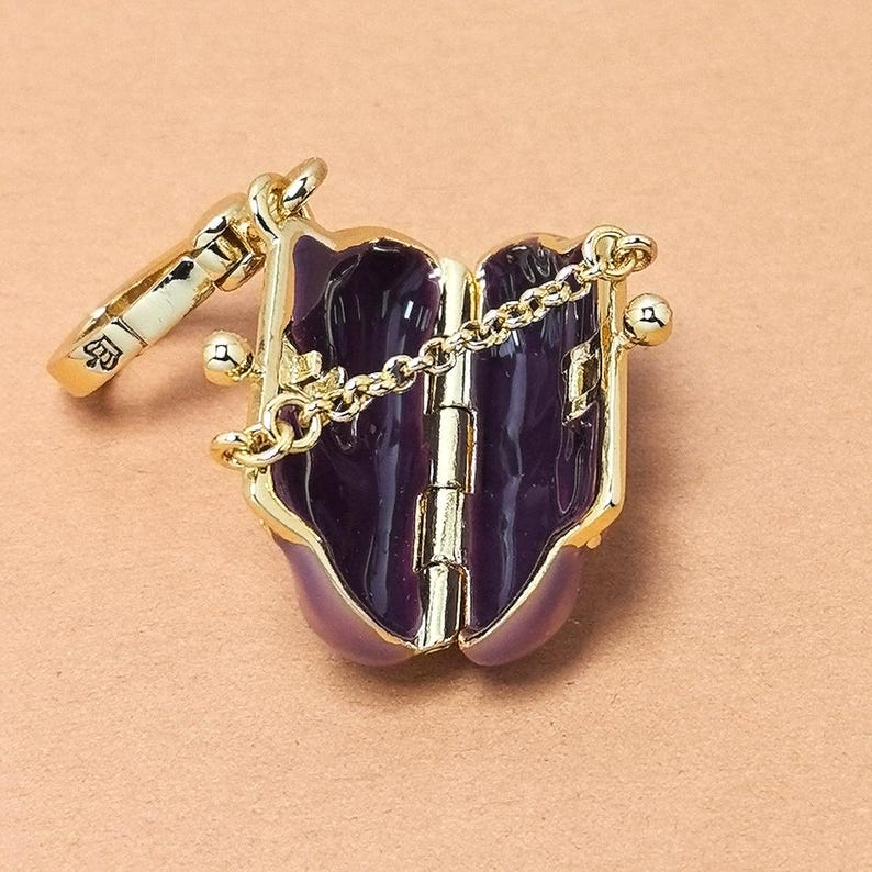 May include: A gold-coloured, hinged locket with a deep purple interior and a delicate chain. The locket is adorned with gold accents and a small, decorative clasp. The design resembles a miniature book or purse.