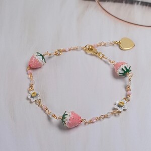 Peut inclure: Un bracelet doré orné de breloques fraises roses avec des détails blancs et des feuilles vertes. Il comprend également de petites fleurs blanches et un charm en forme de cœur. Le bracelet est orné de petites perles roses, pour un style délicat.