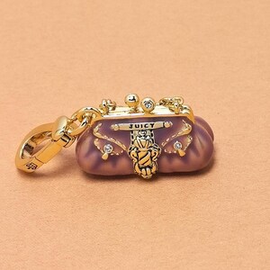 May include: A gold-coloured charm in the shape of a small, purple purse with the word "JUICY" on it. The charm has gold accents, including a clasp, decorative elements, and small rhinestones. The purse charm is a fashion accessory.