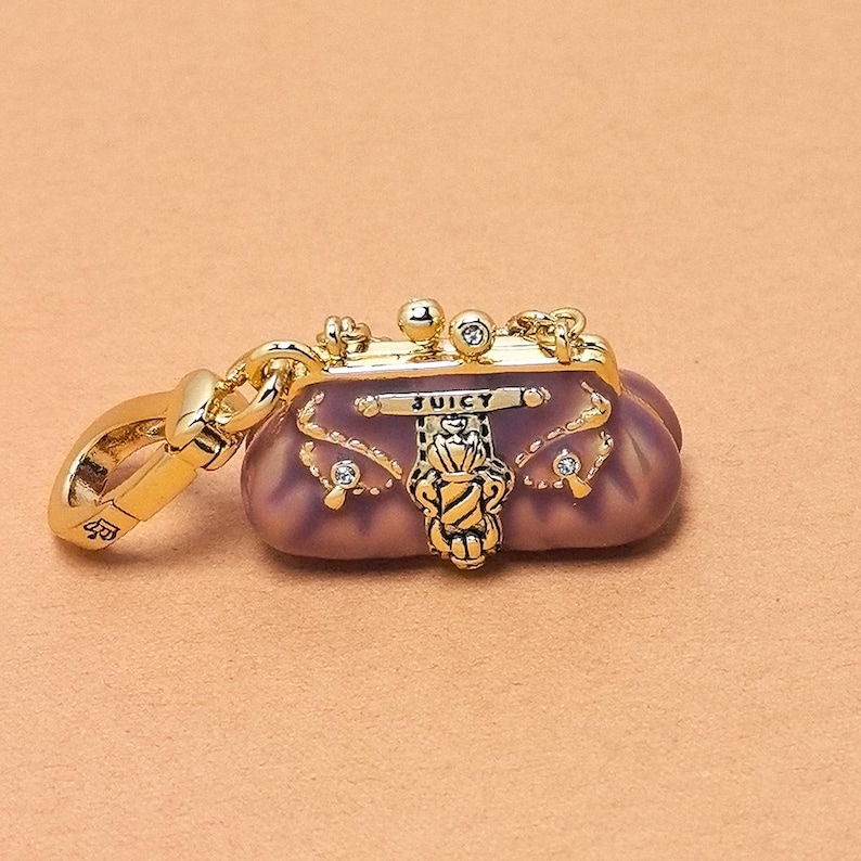 May include: A gold-coloured charm in the shape of a small, purple purse with the word "JUICY" on it. The charm has gold accents, including a clasp, decorative elements, and small rhinestones. The purse charm is a fashion accessory.
