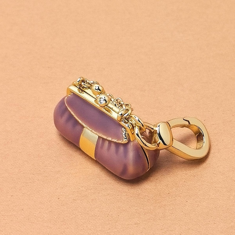 May include: A small purple handbag charm with gold-coloured detailing. The charm has a gold-coloured clasp, chain, and a bar across the top. A gold-coloured stripe is visible on the front of the handbag.