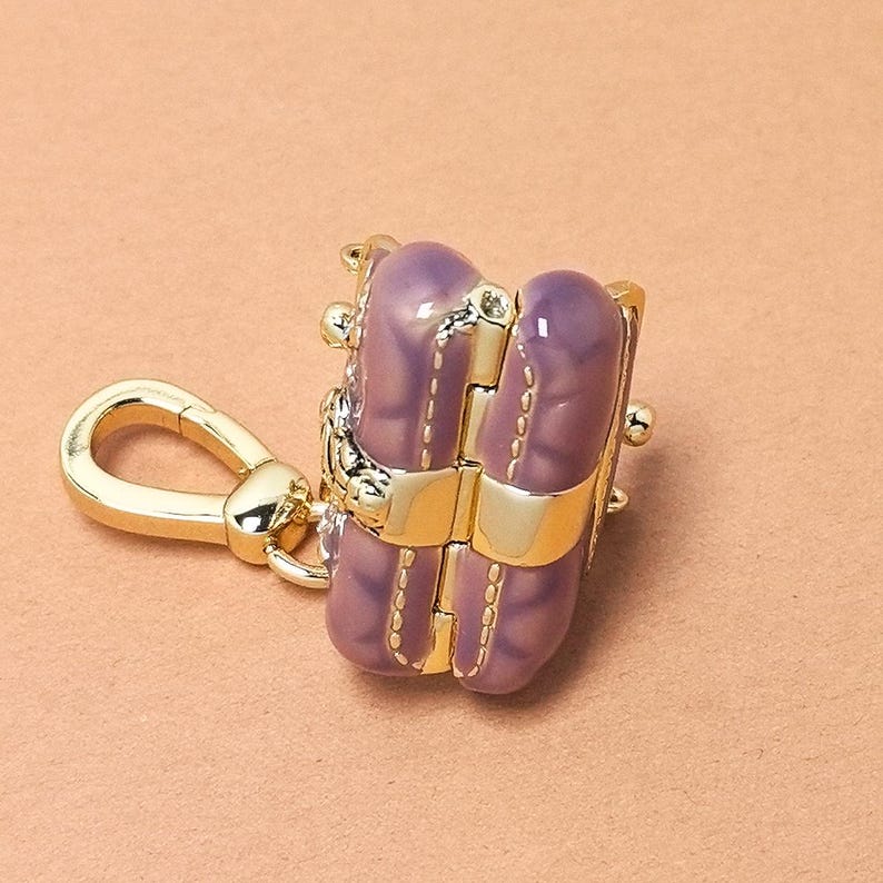 May include: A gold-coloured keychain with a purple and gold charm. The charm is shaped like a small purse with a gold clasp and accents. The purse has a quilted design and a gold clasp. The keychain is on a peach-coloured background.