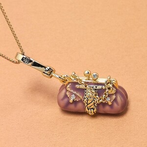 May include: A gold necklace with a small, purple purse pendant. The purse has a clasp closure, a chain, and is decorated with small crystals. The pendant hangs from a delicate gold chain, creating a stylish accessory.