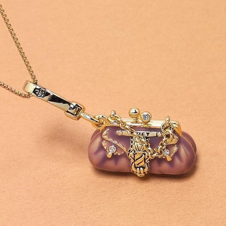 May include: A gold necklace with a small, purple purse pendant. The purse has a clasp closure, a chain, and is decorated with small crystals. The pendant hangs from a delicate gold chain, creating a stylish accessory.