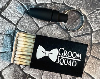 May include: A black matchbox with a white bow tie and the text "Groom Squad". The matchbox is filled with white matches. A black key chain with a silver ring is also in the image.