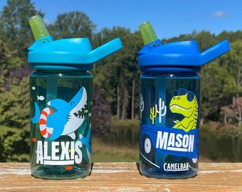 May include: Two blue and green water bottles with straw lids and carrying handles. The bottle on the left has a shark graphic and the name "ALEXIS" printed on it. The bottle on the right has a dinosaur graphic and the name "MASON" printed on it. Both bottles have the brand name "CAMELBAK" printed on them.