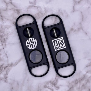 May include: Two black cigar cutters with a silver blade. One cutter has the initials "SSM" in a circle and the other has the initials "ADS" in a rectangle.