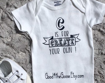 May include: White baby onesie with black text that reads "C is for CREATE YOUR OWN!" and a website address. The onesie is on a gray fuzzy surface with a pair of white baby shoes.