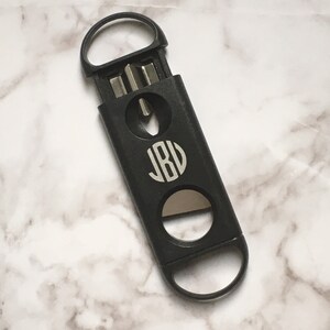 May include: Black cigar cutter with a monogrammed circle that says "JBV" on the front. The cutter has a ring on the top and a blade on the bottom.