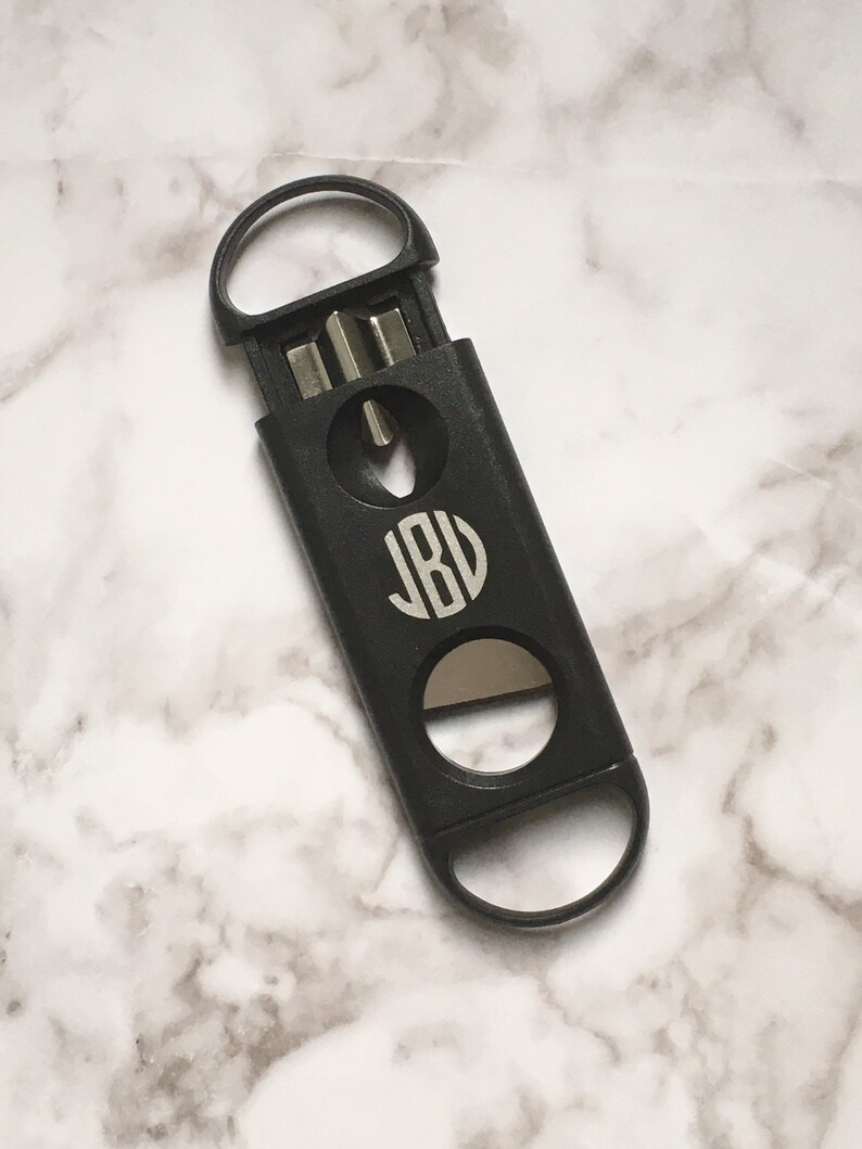 May include: Black cigar cutter with a monogrammed circle that says "JBV" on the front. The cutter has a ring on the top and a blade on the bottom.