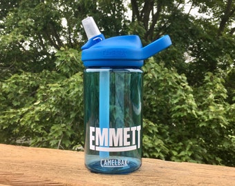 May include: Blue water bottle with a straw and a blue lid. The bottle has a white label with the name "EMMETT" and the brand name "CAMELBAK" printed on it.