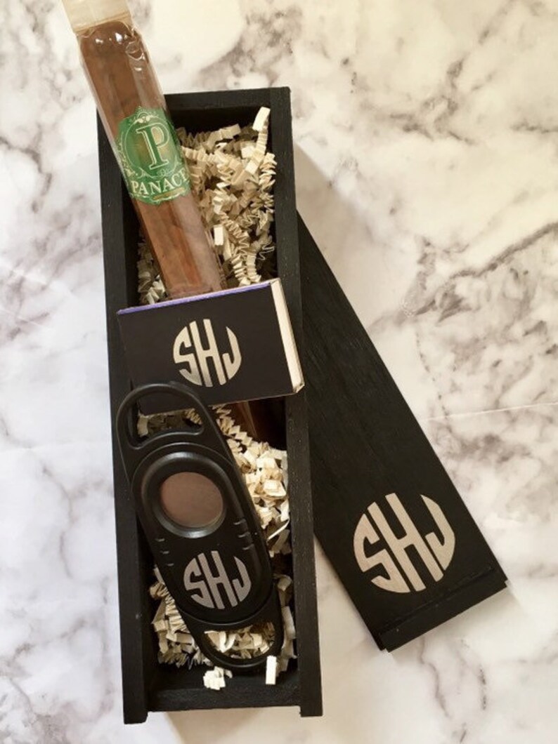 May include: A black wooden box with a cigar, a cigar cutter, and a matchbook. The box is decorated with the initials "SHJ".