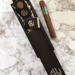 May include: A black wooden box with a cigar cutter, matches, and a cigar. The box is engraved with the initials "SHJ".