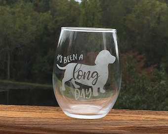 May include: A clear glass wine glass with the text "It's been a long day" and a silhouette of a dachshund etched on the side.