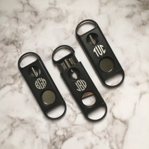 May include: Three black cigar cutters with different monogrammed initials. Each cutter has a silver blade and a round opening for cutting cigars.