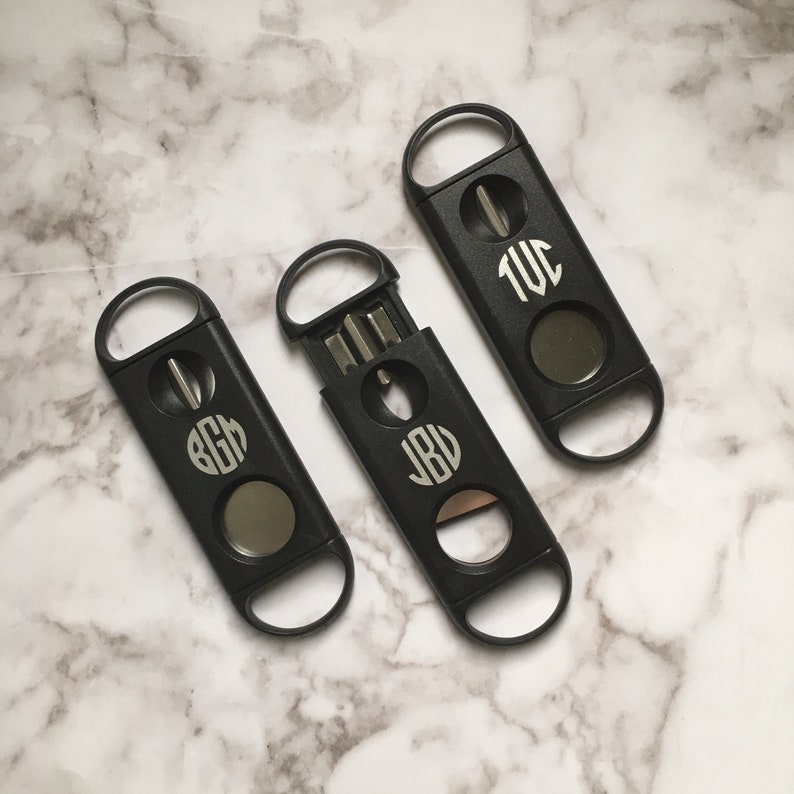 May include: Three black cigar cutters with different monogrammed initials. Each cutter has a silver blade and a round opening for cutting cigars.