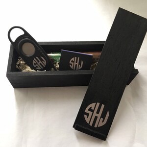 May include: A black wooden box with a lid, containing a cigar cutter, a cigar, and a matchbook. The box and the items inside are monogrammed with the initials "SHJ".