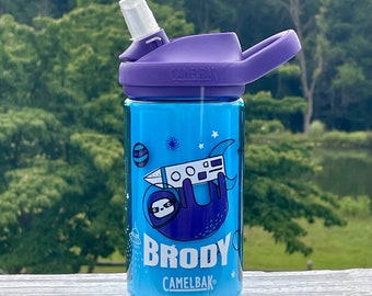 May include: A blue water bottle with a purple lid and a straw. The bottle has a space-themed design with a sloth riding a rocket. The bottle says "Brody" and "CamelBak" on it.