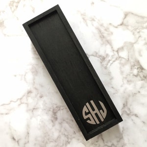 May include: A black wooden box with a raised, circular, silver monogram of the letters "SHJ".