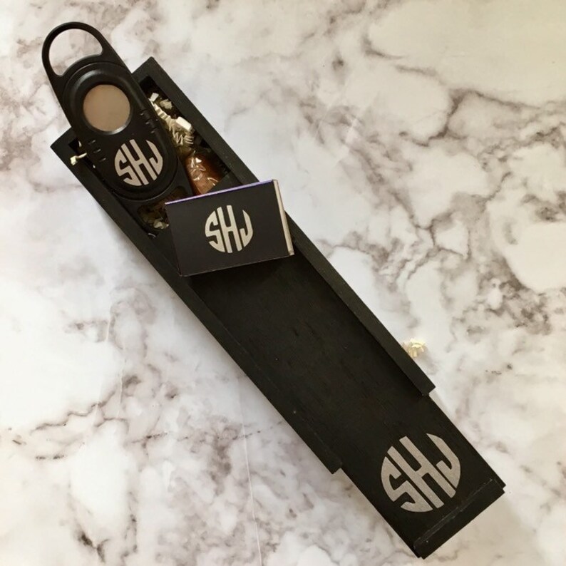 May include: A black wooden box with a cigar cutter, a cigar, and a matchbook. The box is engraved with the initials "SHJ".