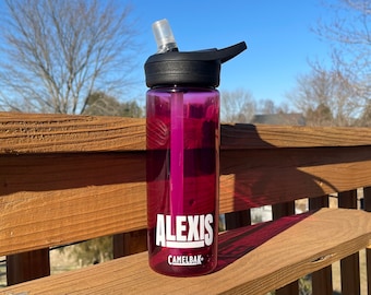 Puede incluir: Una botella de agua rosa con tapa negra y una pajita. La botella tiene el nombre "Alexis" y la marca "CamelBak" impresa en ella.