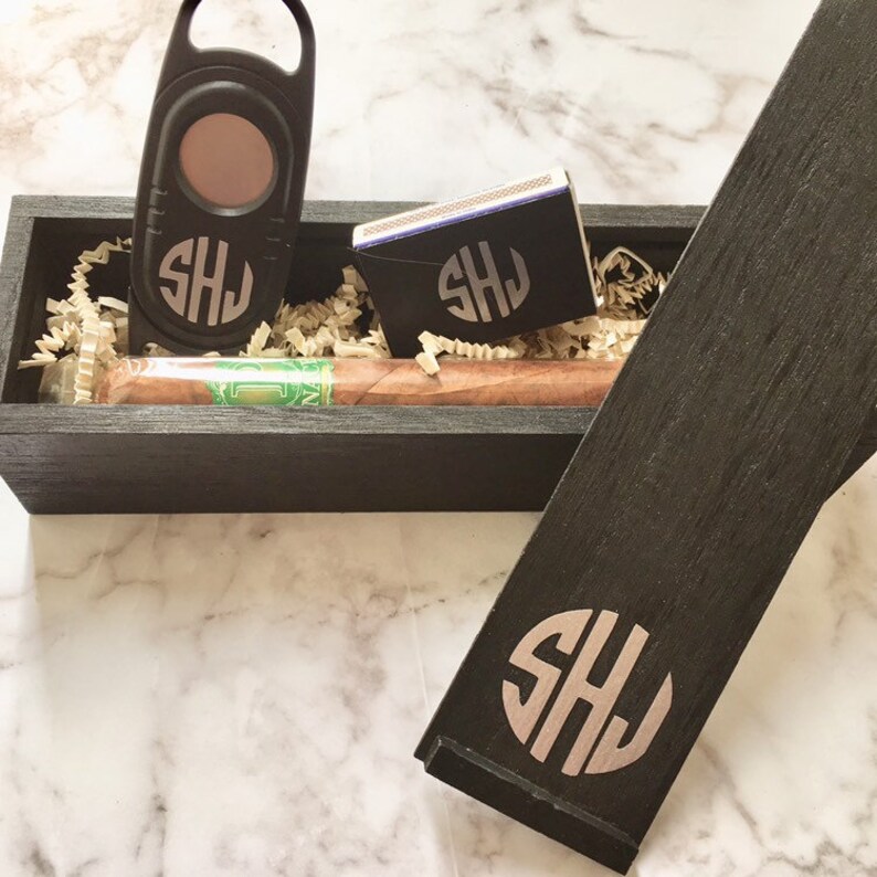 May include: A black wooden box with a cigar, a cigar cutter, and a matchbook. The box and the accessories are engraved with the initials "SHJ".