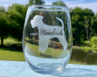 May include: A clear stemless wine glass with the words "I HAVE Standards" and a dog silhouette etched on the front. The glass is sitting on a light blue surface with a blurred green background.