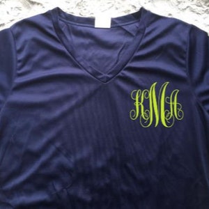 Puede incluir: Una camiseta azul marino de manga corta con cuello en V y un monograma verde de las letras "KMA" en la parte delantera.
