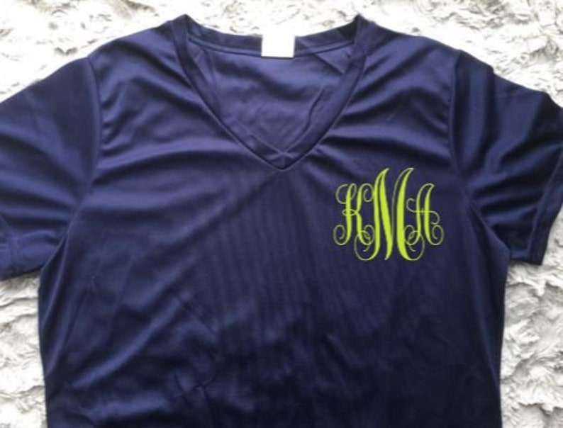 Puede incluir: Una camiseta azul marino de manga corta con cuello en V y un monograma verde de las letras "KMA" en la parte delantera.
