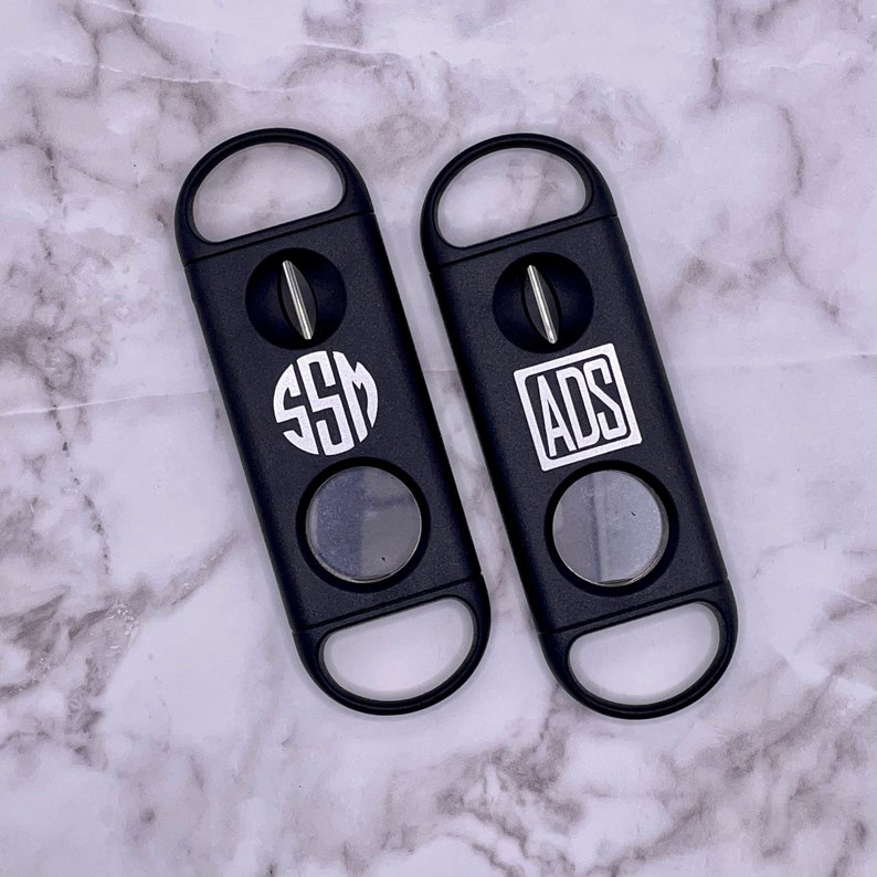 May include: Two black cigar cutters with a silver blade. One cutter has the initials "SSM" in a circle and the other has the initials "ADS" in a rectangle.