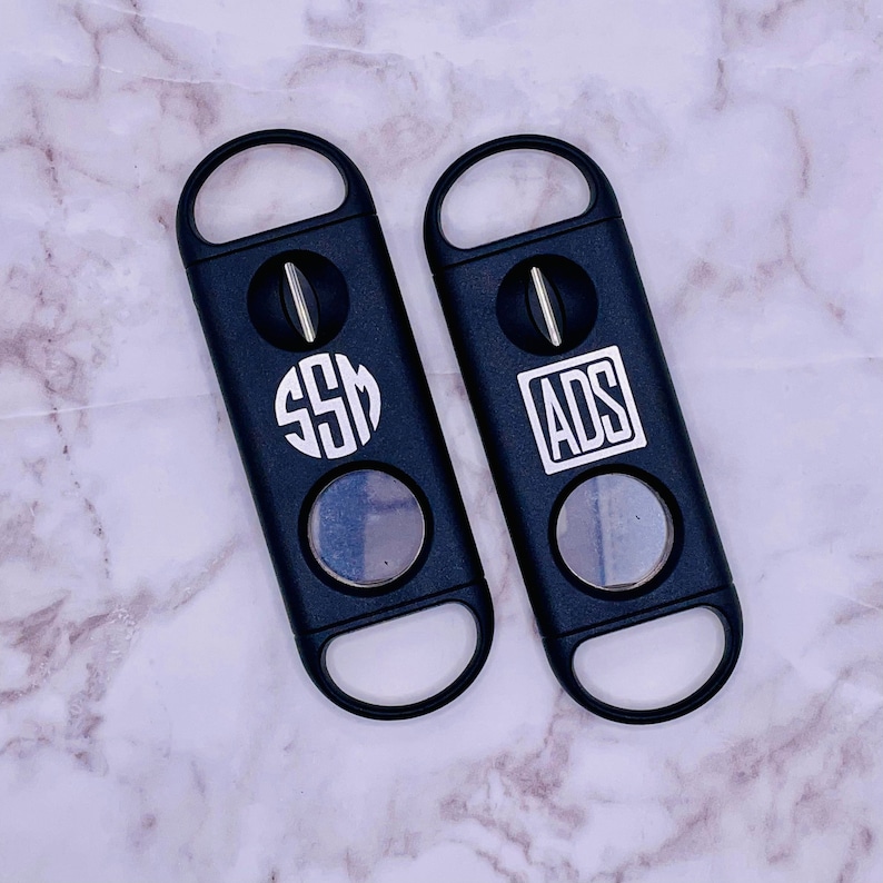 May include: Two black cigar cutters with a monogrammed design. The cutter on the left has the monogram "SSM" and the cutter on the right has the monogram "ADS".