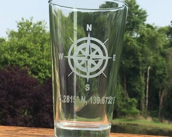 May include: A clear glass pint glass with a compass and coordinates etched on the side. The coordinates are 5.2815 N, 139.6721.