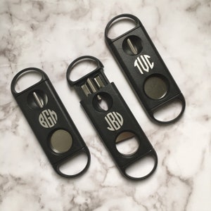 May include: Three black cigar cutters with monogrammed initials. The cutters have a round blade and a small hole for pushing out the cut end of the cigar. The initials are "J.B.V.", "B.G.H.", and "T.W.C."