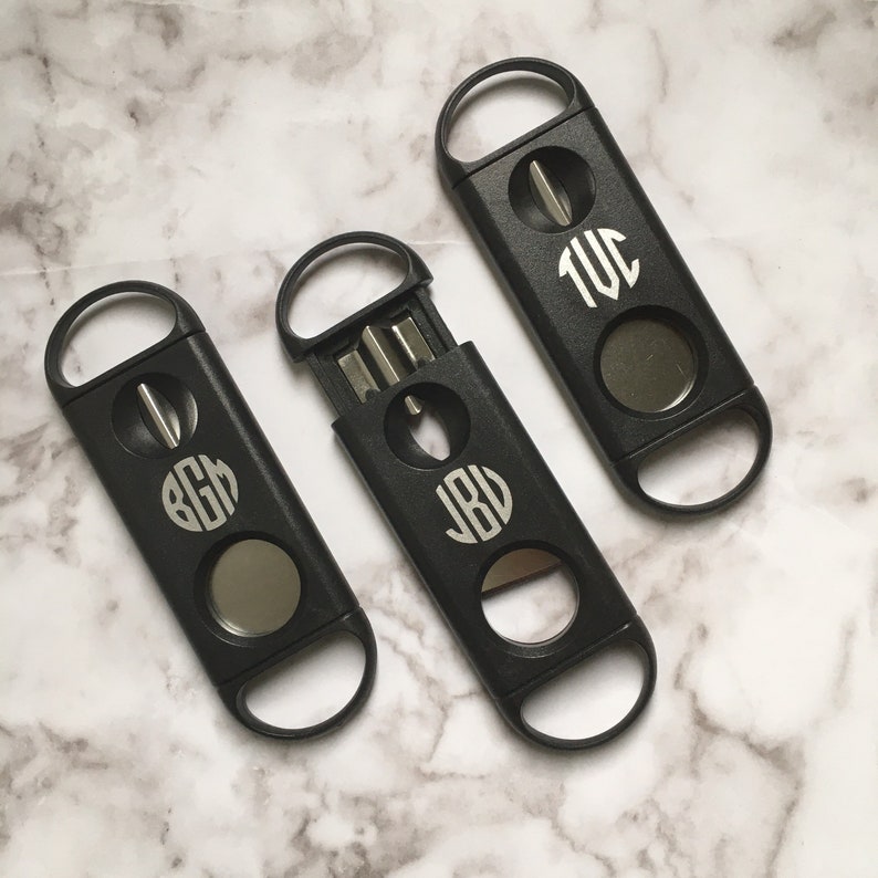 May include: Three black cigar cutters with monogrammed initials. The cutters have a round blade and a small hole for pushing out the cut end of the cigar. The initials are "J.B.V.", "B.G.H.", and "T.W.C."