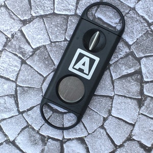 May include: Black cigar cutter with a silver blade and a white "A" logo. The cutter is on a grey stone background.